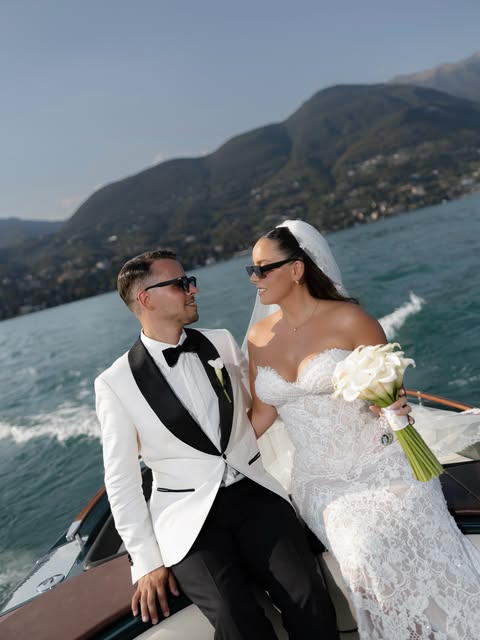 A couple in wedding attire on a boat with mountains and a lake behind them