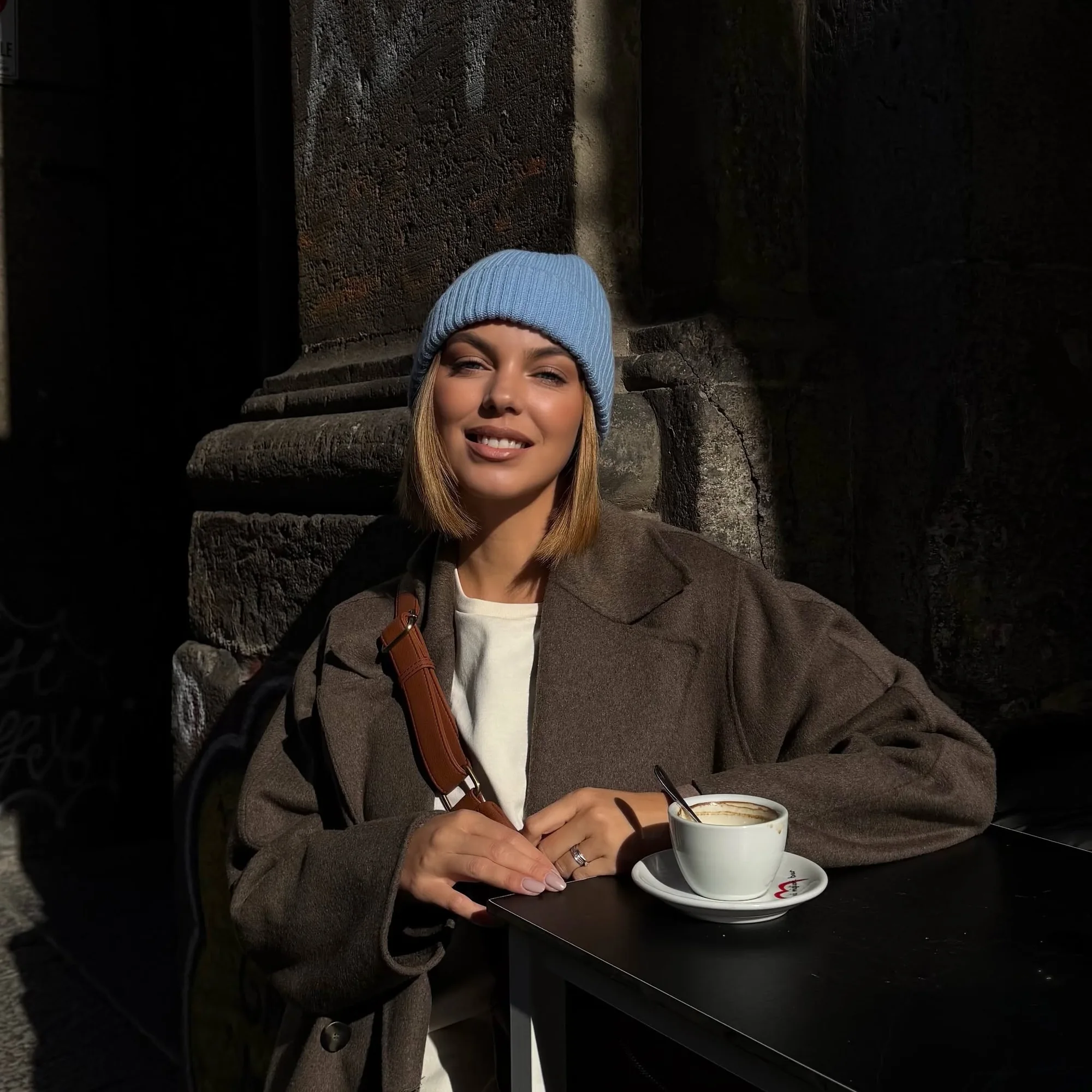 Carly smiling at a café table in sunlight, wearing a blue beanie