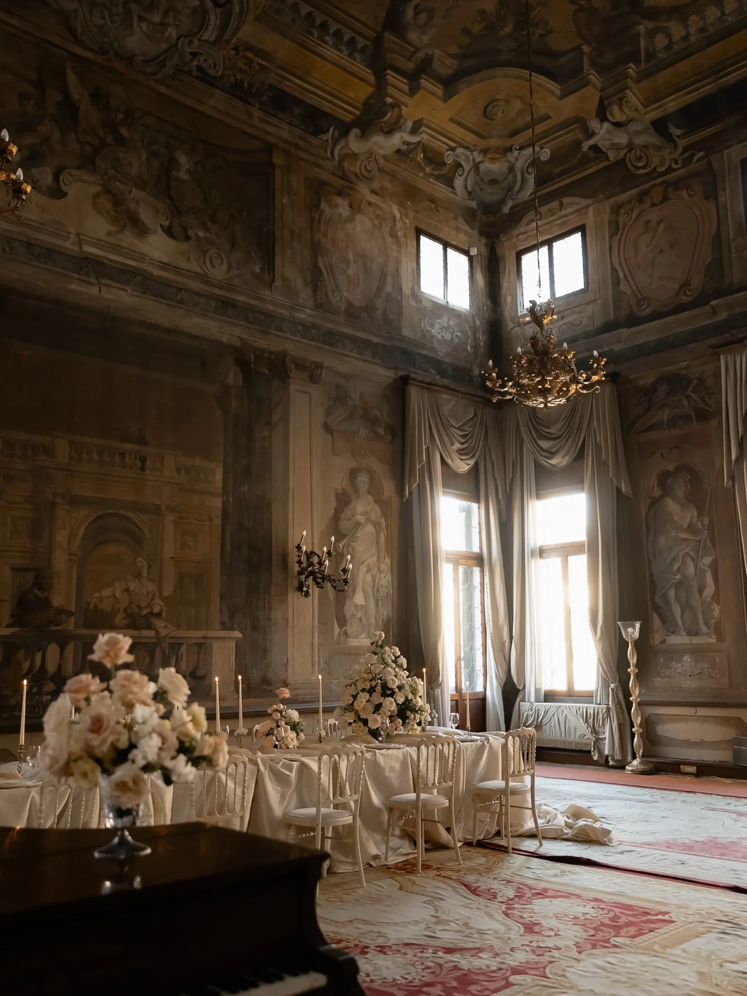 An ornate palazzo reception room with chandeliers, flowers, candlelit tables, and a grand piano