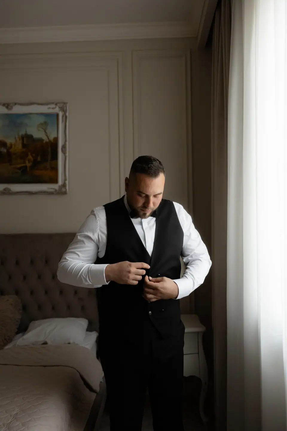 Norbert buttoning his black waistcoat by the bedroom window