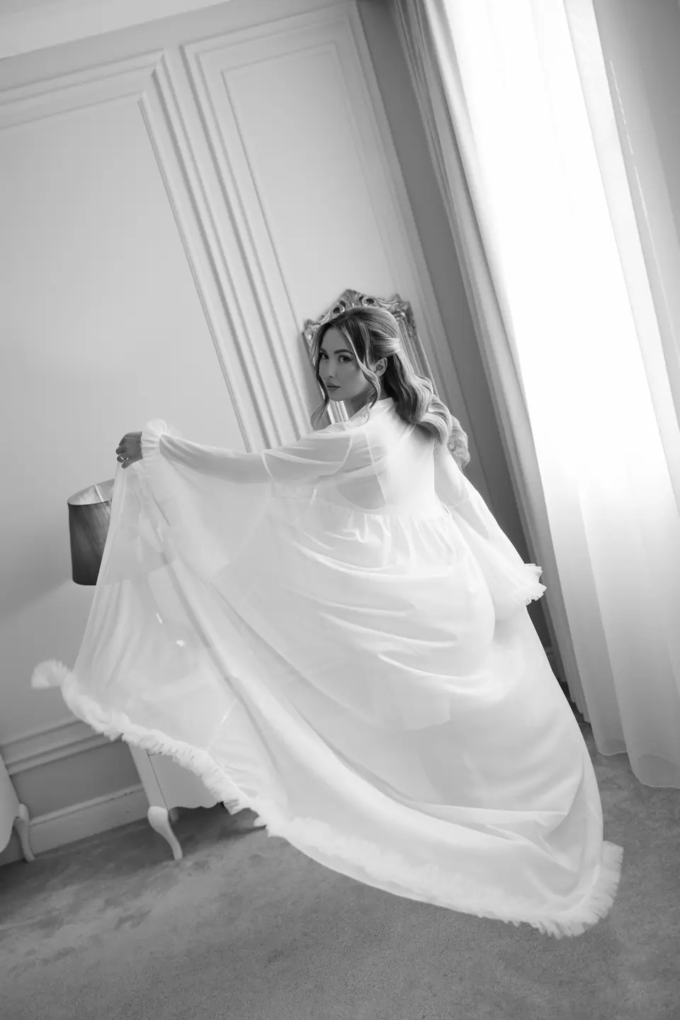 Alexandra in black and white lifting her sheer robe by the window