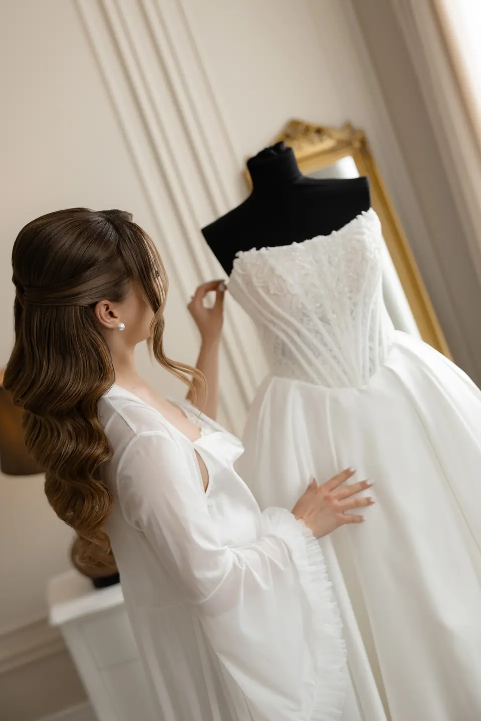Alexandra in her robe touching the bodice of her wedding dress on the mannequin