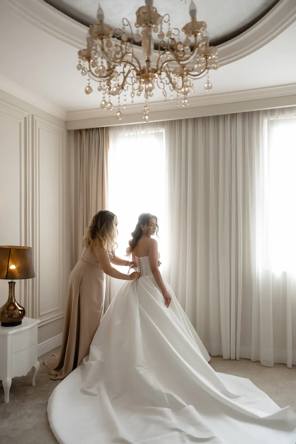 A bridesmaid fastening the back of Alexandra’s dress beneath a chandelier