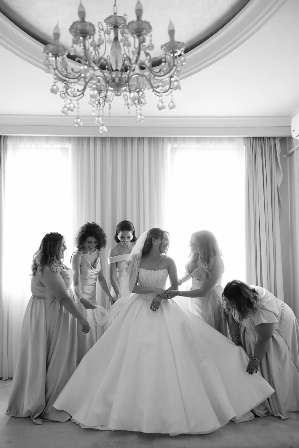 Alexandra in her gown surrounded by bridesmaids beneath a chandelier