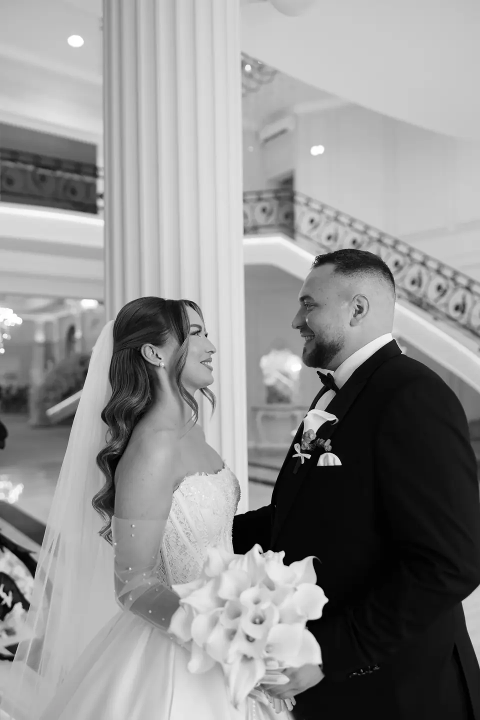 Alexandra and Norbert face to face by a white column, her calla lily bouquet in hand
