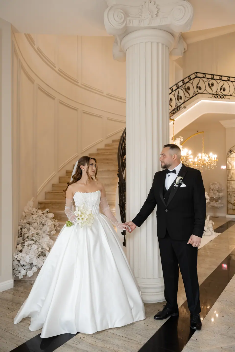 Alexandra and Norbert holding hands by a column at the foot of a curved staircase