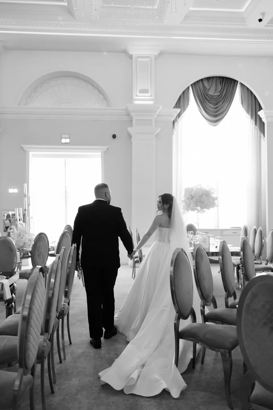 Alexandra and Norbert walking hand in hand between the reception chairs, in black and white