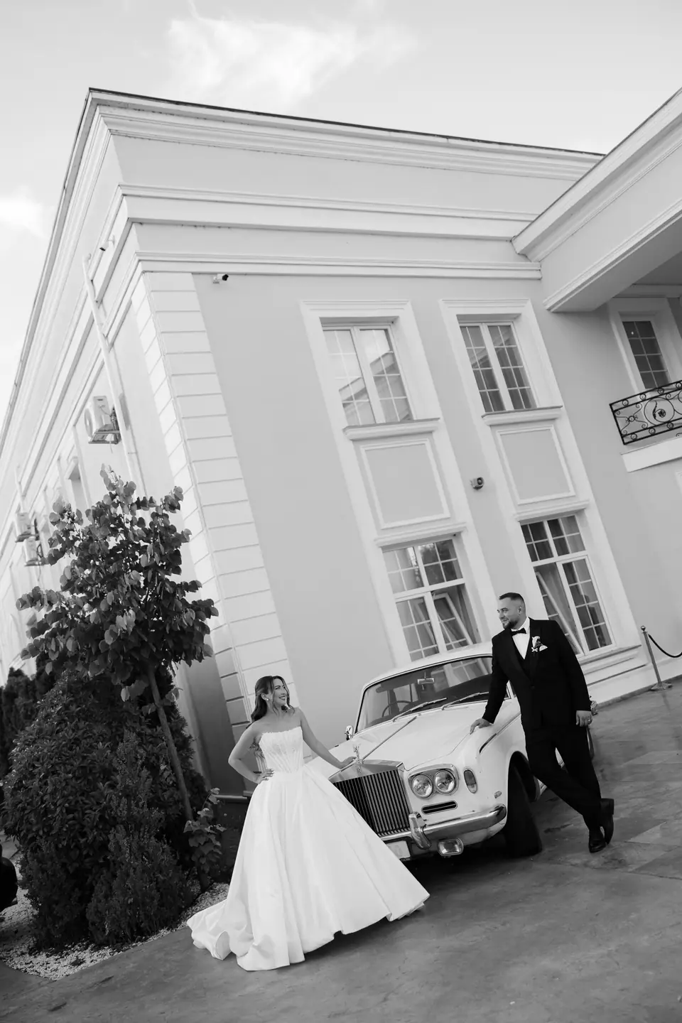 Alexandra and Norbert beside a vintage white car outside the venue, in black and white