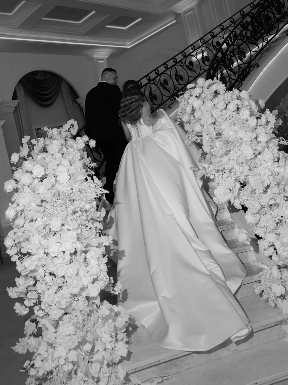 Alexandra in her gown on the staircase among white flower arrangements, in black and white