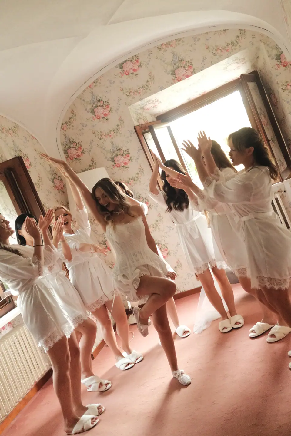 Ana and her bridesmaids in white robes dancing in a room with floral wallpaper