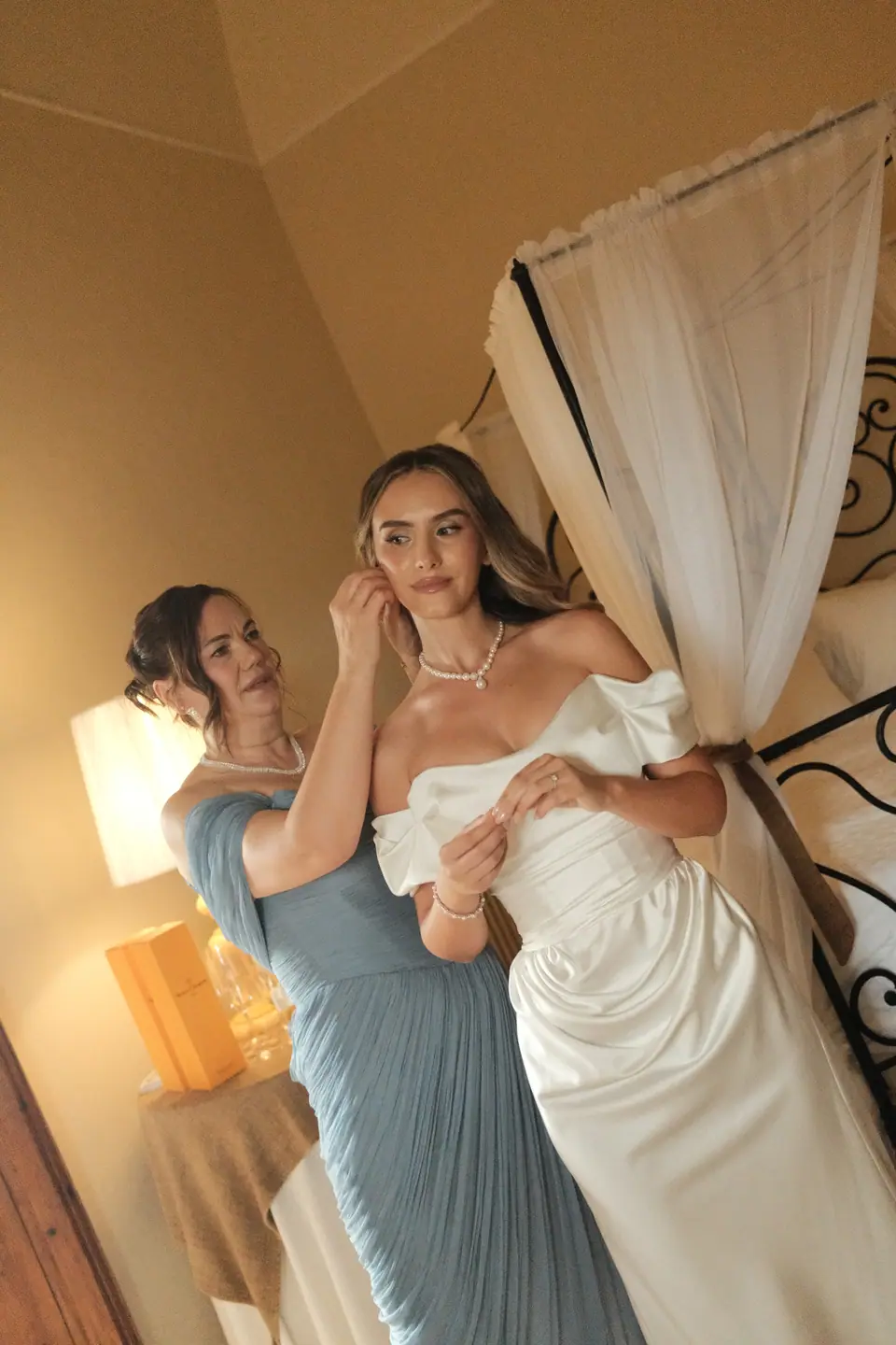 A bridesmaid in pale blue fastening Ana’s earring beside the canopy bed
