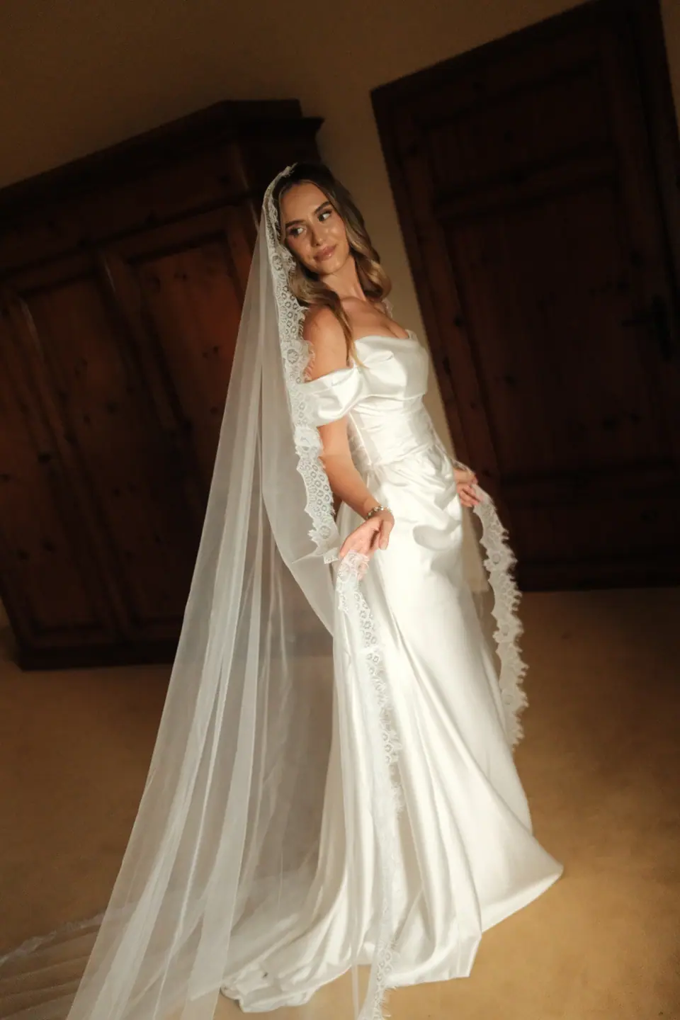 Ana in her off-the-shoulder satin gown and lace veil between dark wooden doors