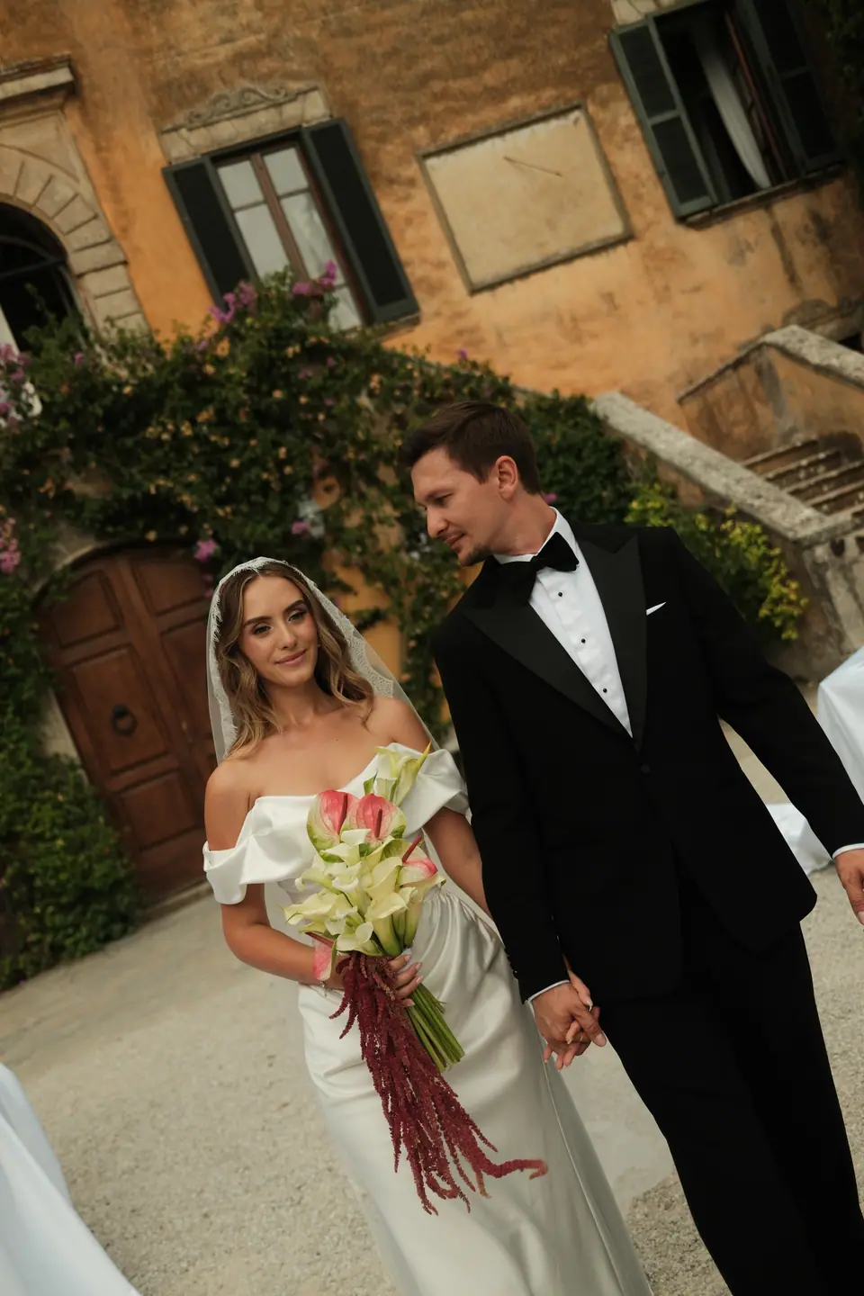 Ana holding a bouquet of white and red flowers beside Silviu in front of the villa