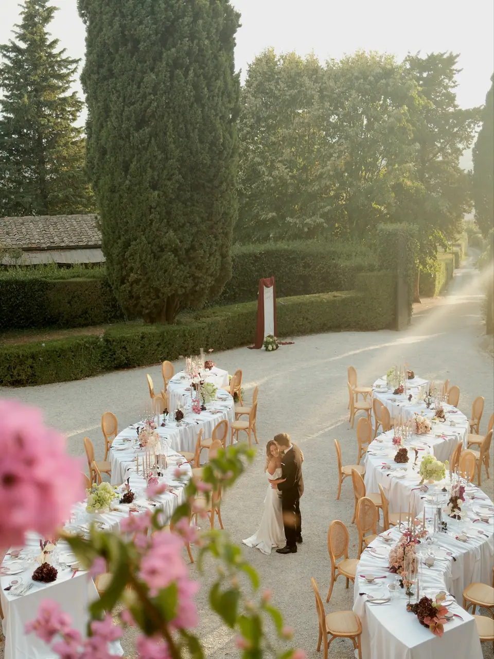 Ana and Silviu kissing among the courtyard tables, seen through pink flowers