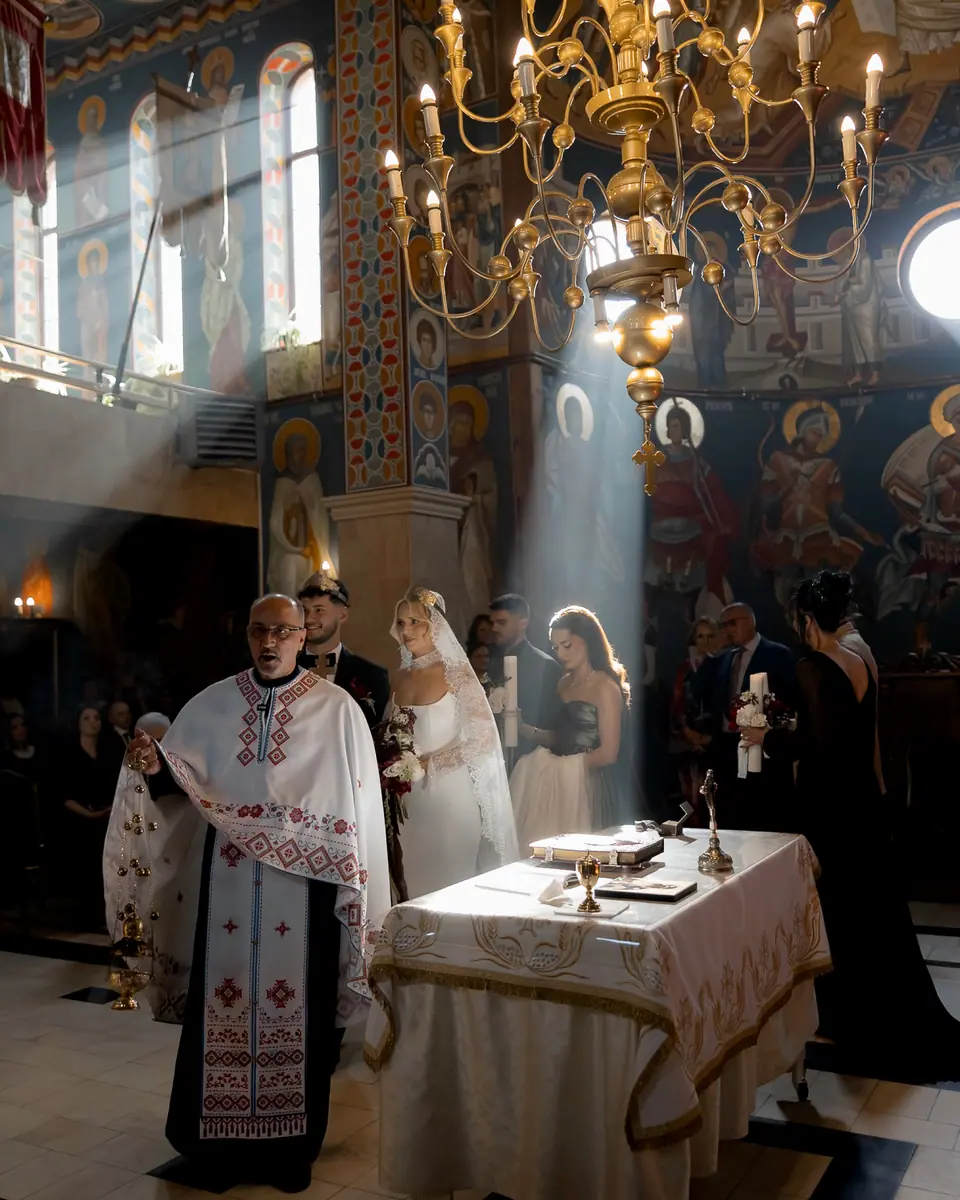 The priest leading the Orthodox ceremony beneath a chandelier, light falling through the church