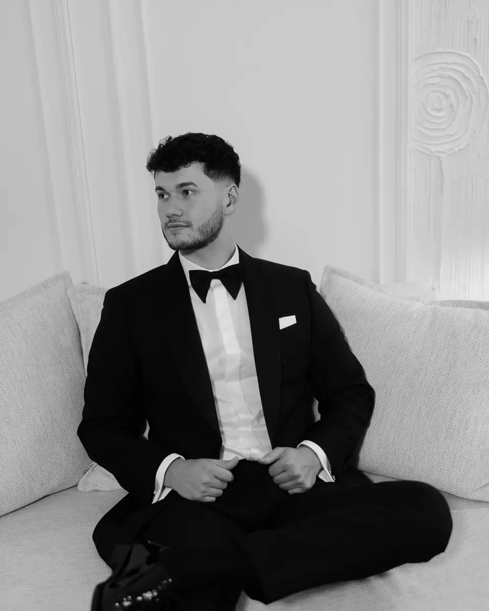 A black and white portrait of Bogdan in a tuxedo sitting on a white sofa