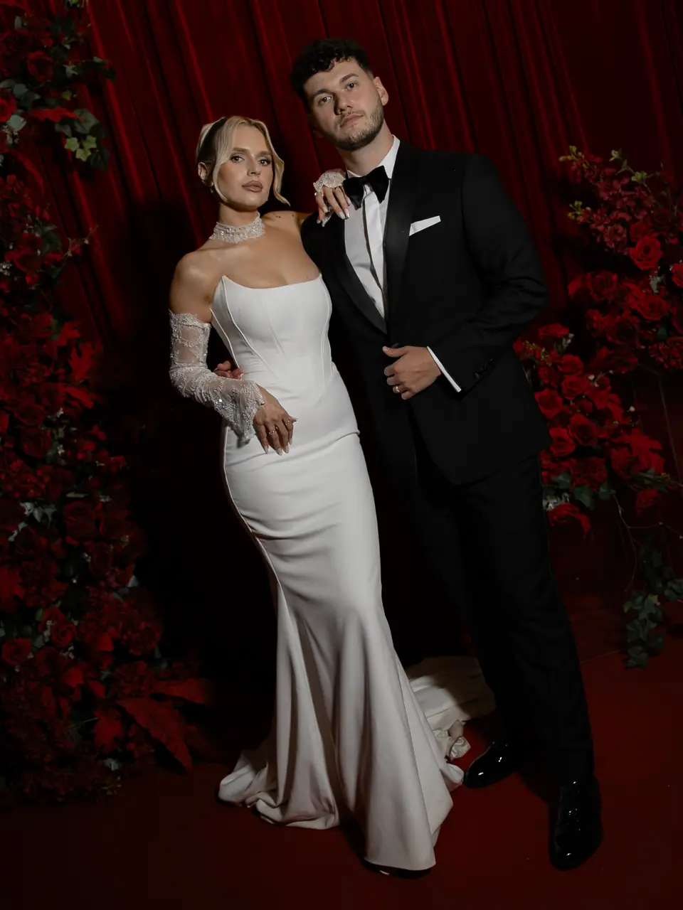 Andreea and Bogdan standing together before deep red drapes and roses