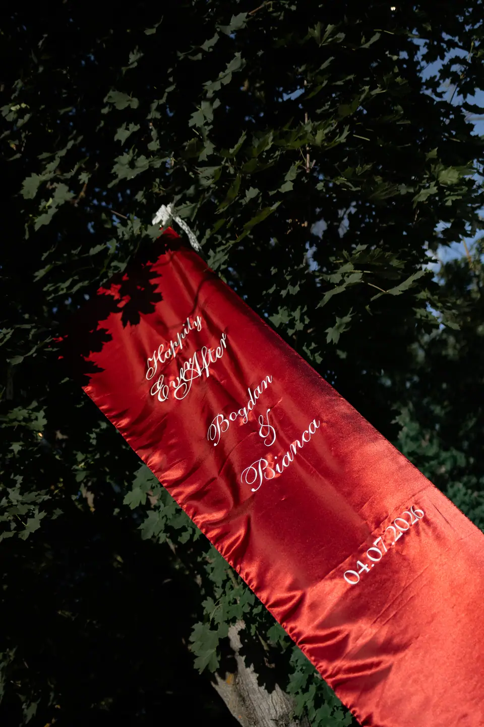 A red satin banner reading “Happily Ever After, Bogdan & Bianca, 04.07.2026” hanging in a tree