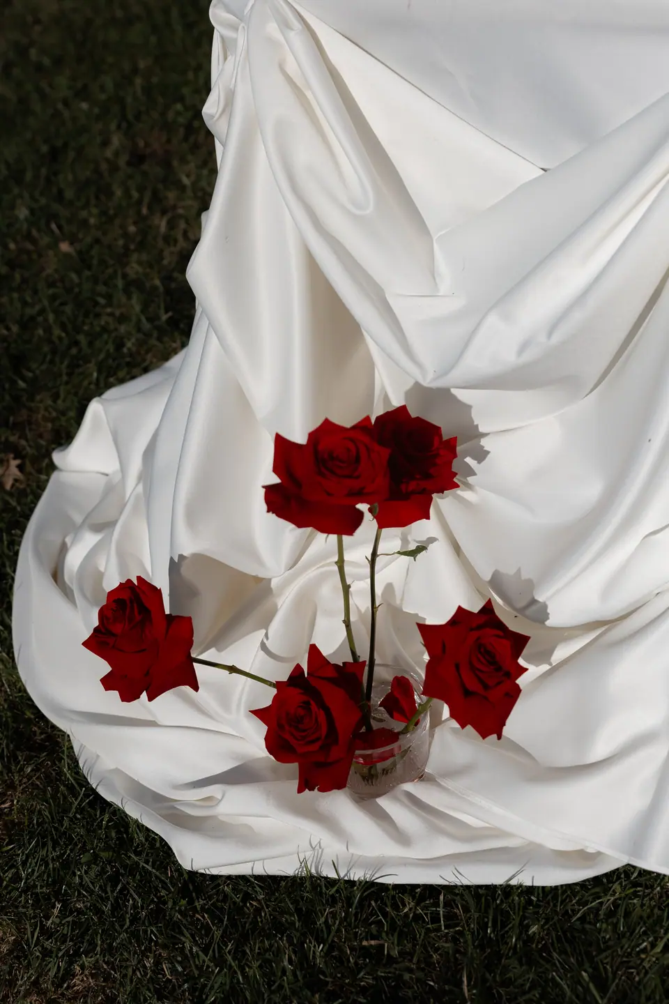 Red roses in a glass on white draped fabric spilling onto the grass