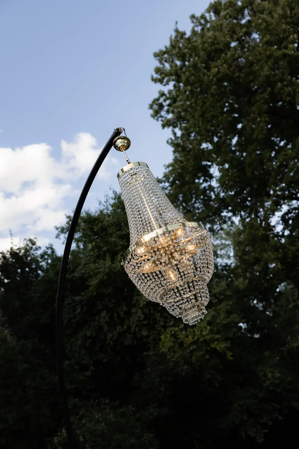 A crystal chandelier hanging from a curved iron stand against the trees