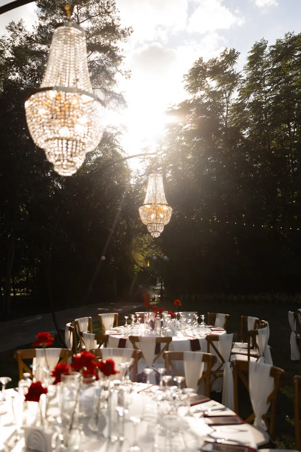 Sunlight through the trees and a chandelier above tables set with red roses
