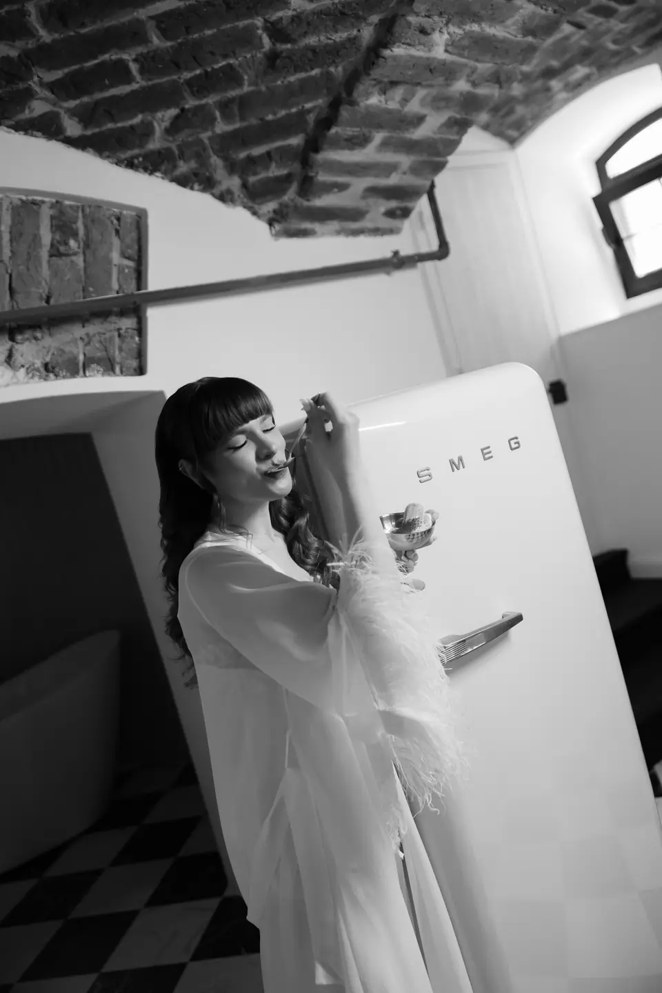 Black and white photograph of Bianca in a feathered robe sipping by the Smeg fridge