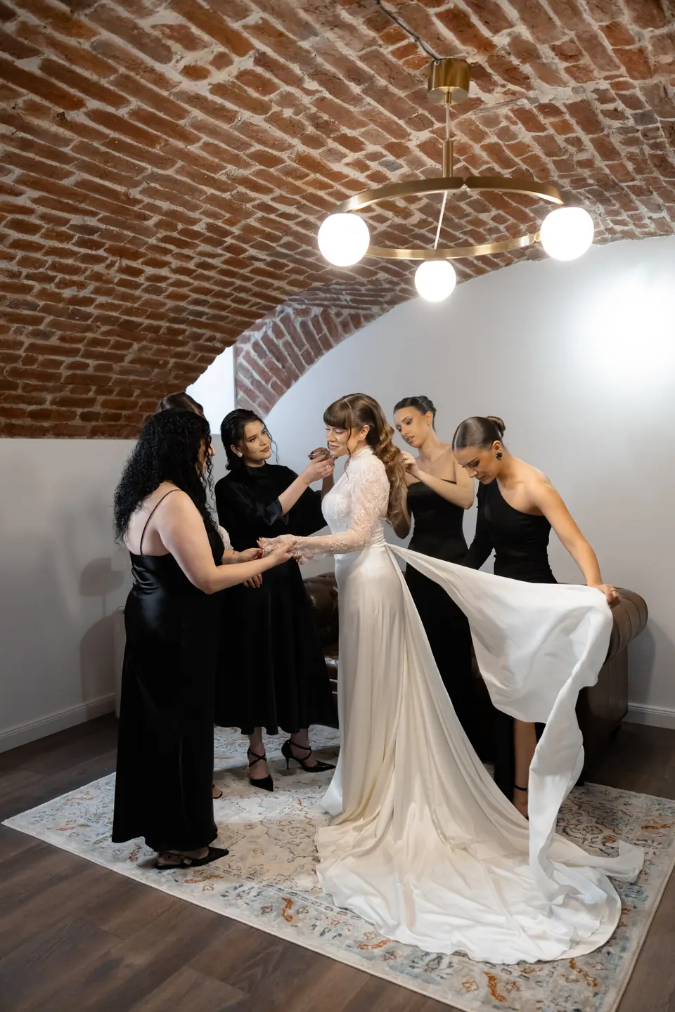 Bridesmaids in black helping Bianca into her gown in a brick-vaulted room