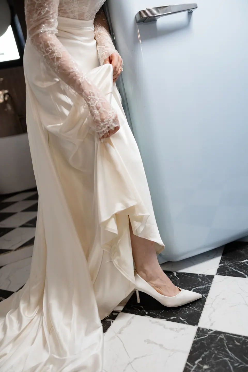 Bianca lifting her gown to show white heels on a black and white floor by the blue fridge