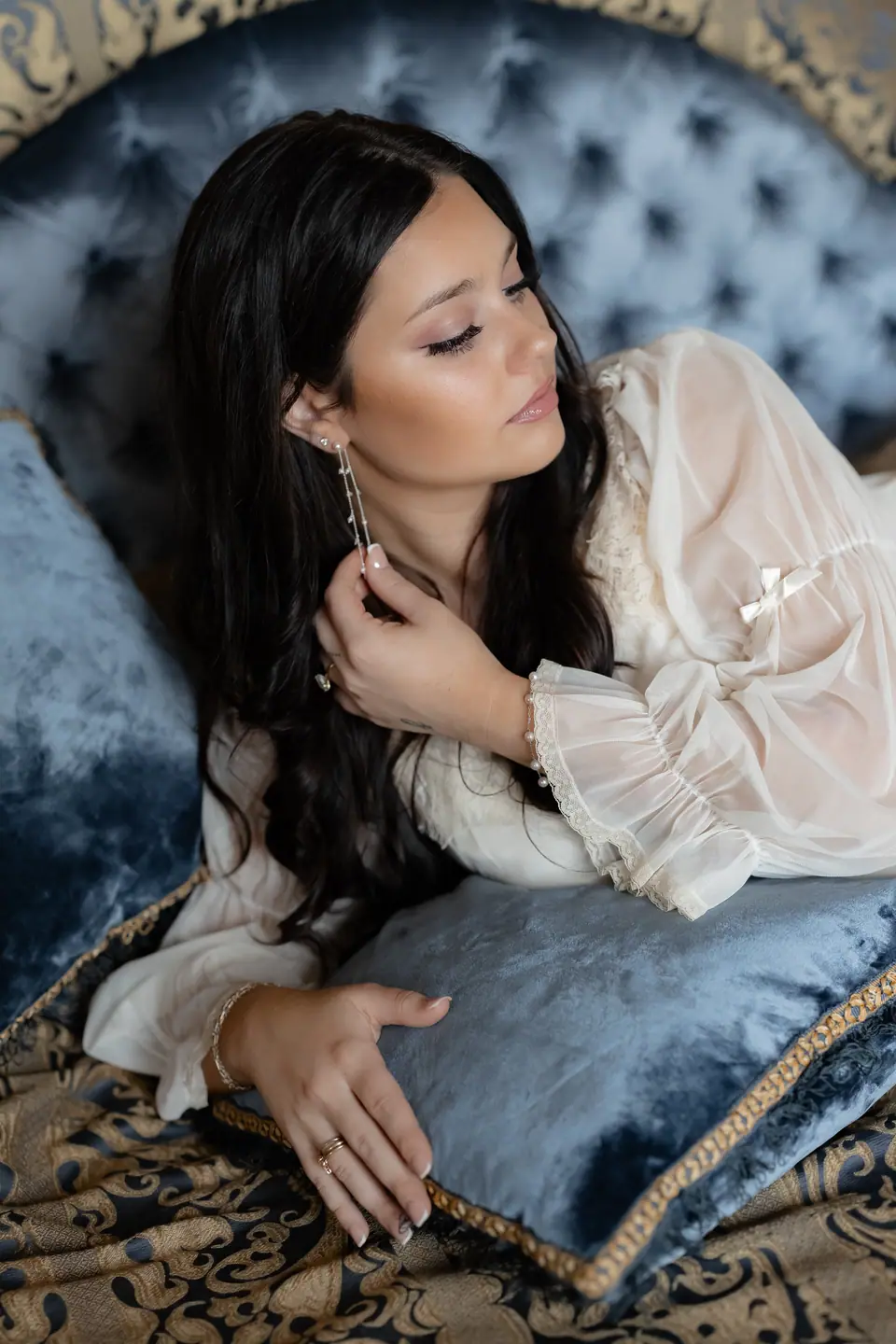 Haley resting on a blue velvet chaise in a sheer robe, putting on an earring