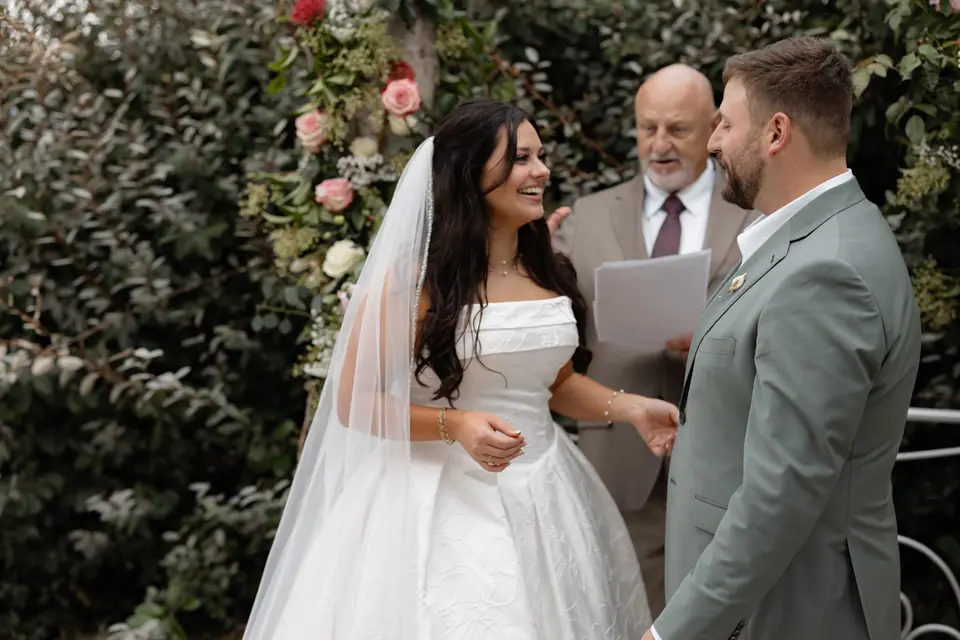 Haley and Zach laughing with the officiant beside a rose-covered hedge during the ceremony