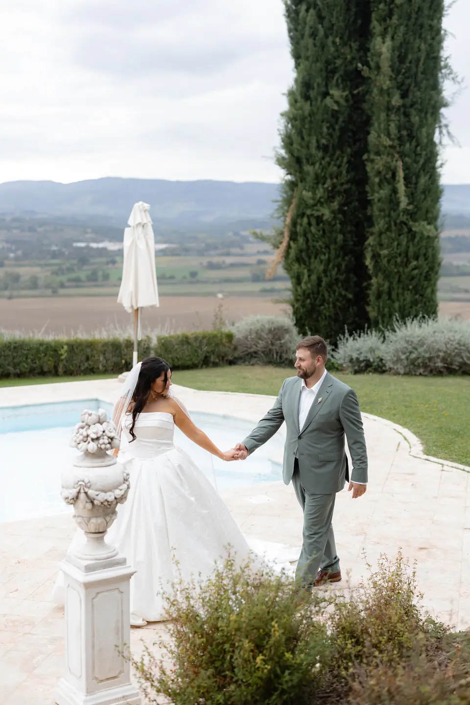 Haley and Zach walking hand in hand past the pool, with a cypress and the Umbrian valley behind