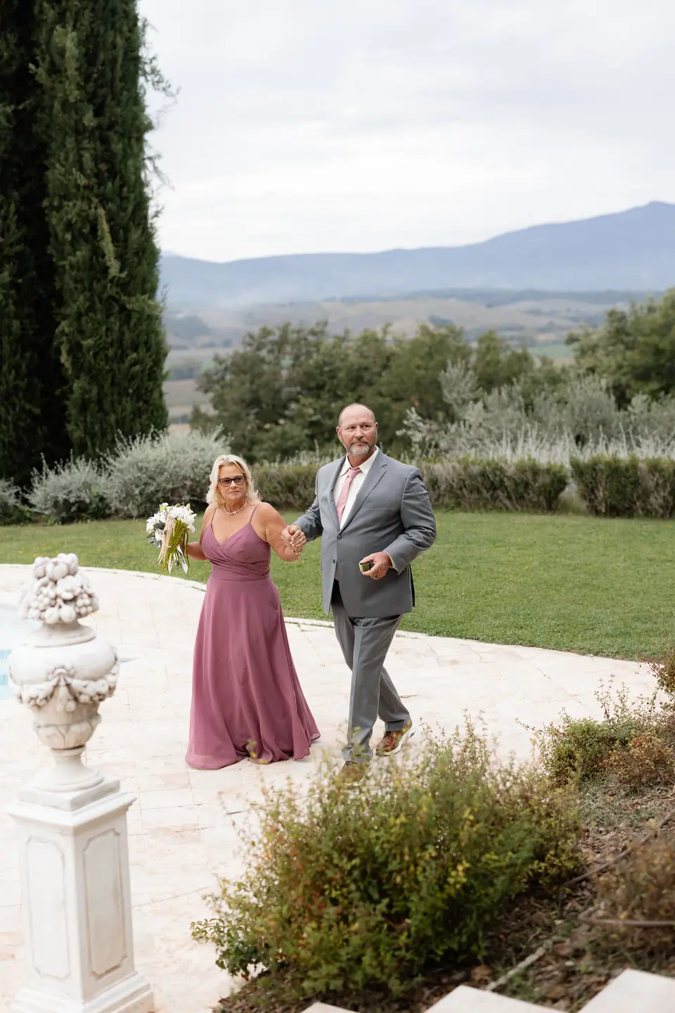 A woman in mauve and a man in grey walking hand in hand by the pool, cypresses and hills behind