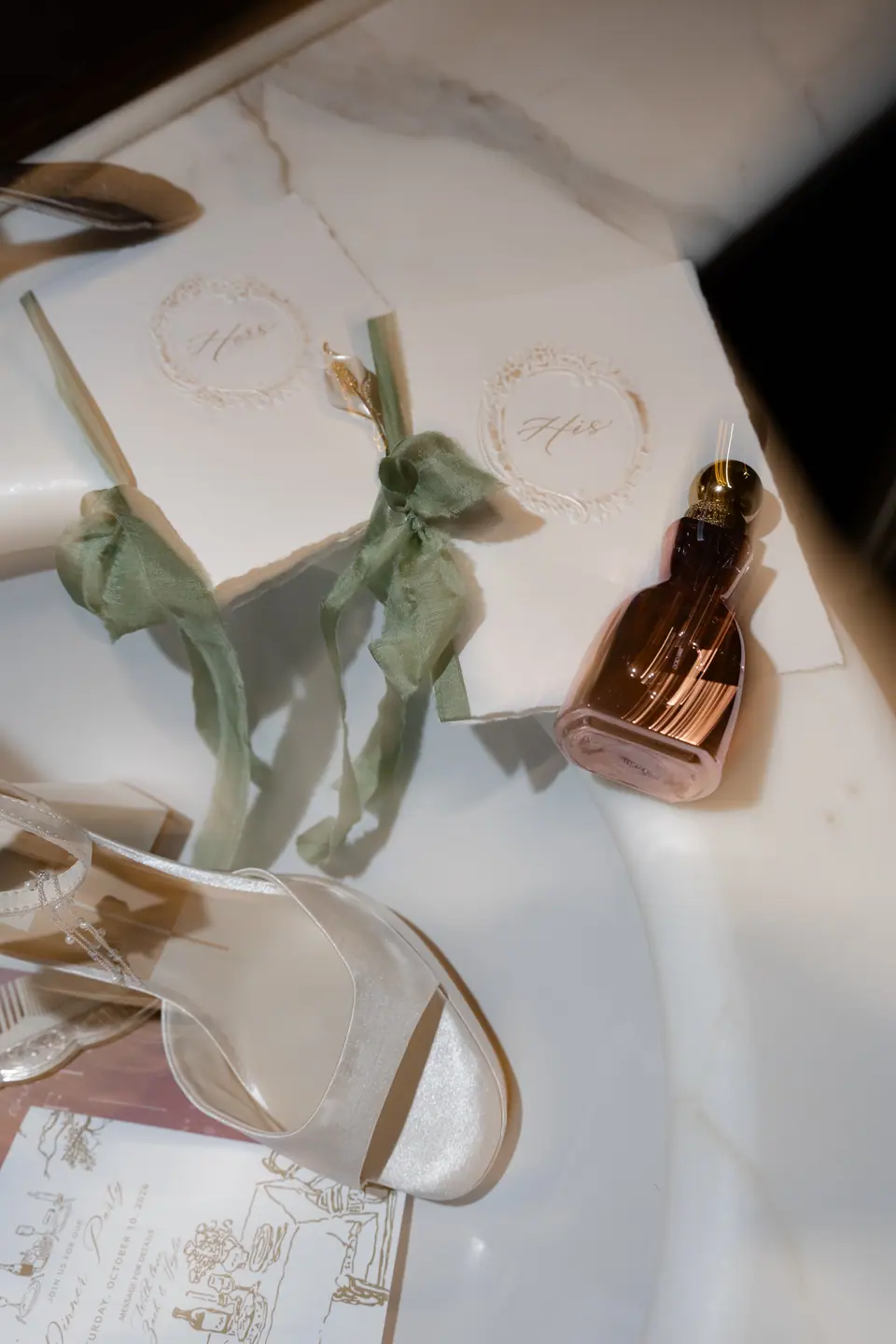 Calligraphy vow books tied with green ribbon beside perfume and bridal shoes on marble