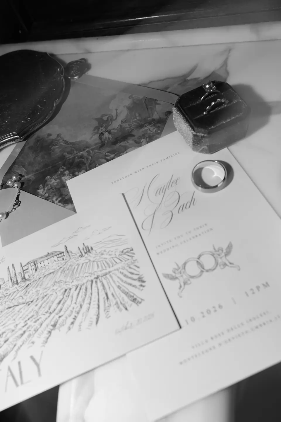 Black and white flat lay of the invitation, a ring box and two rings on a hand-drawn villa map