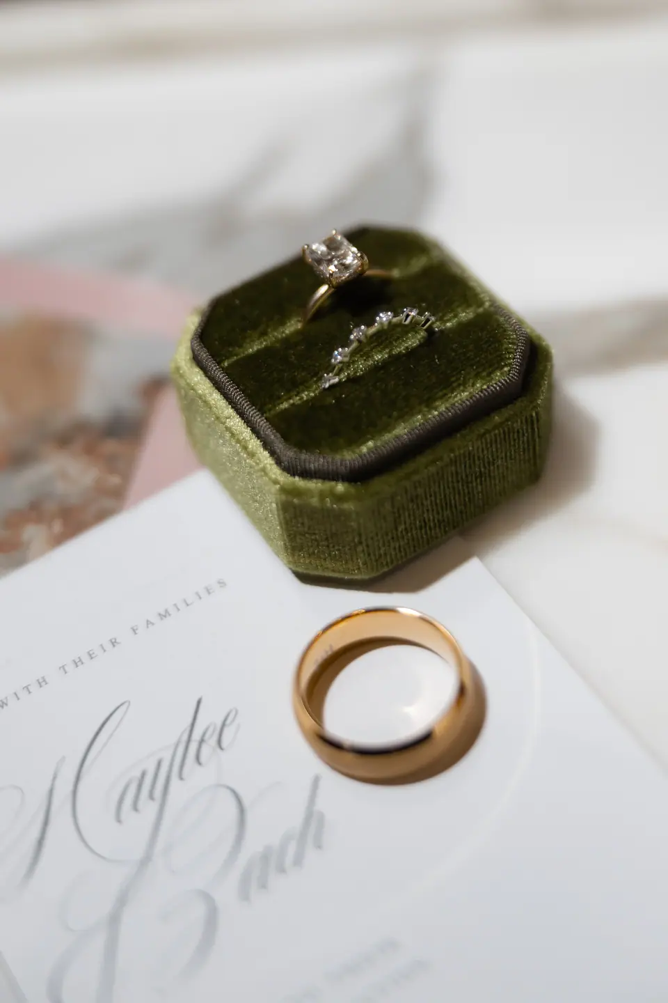 An engagement ring in a green velvet box beside a gold wedding band on the invitation