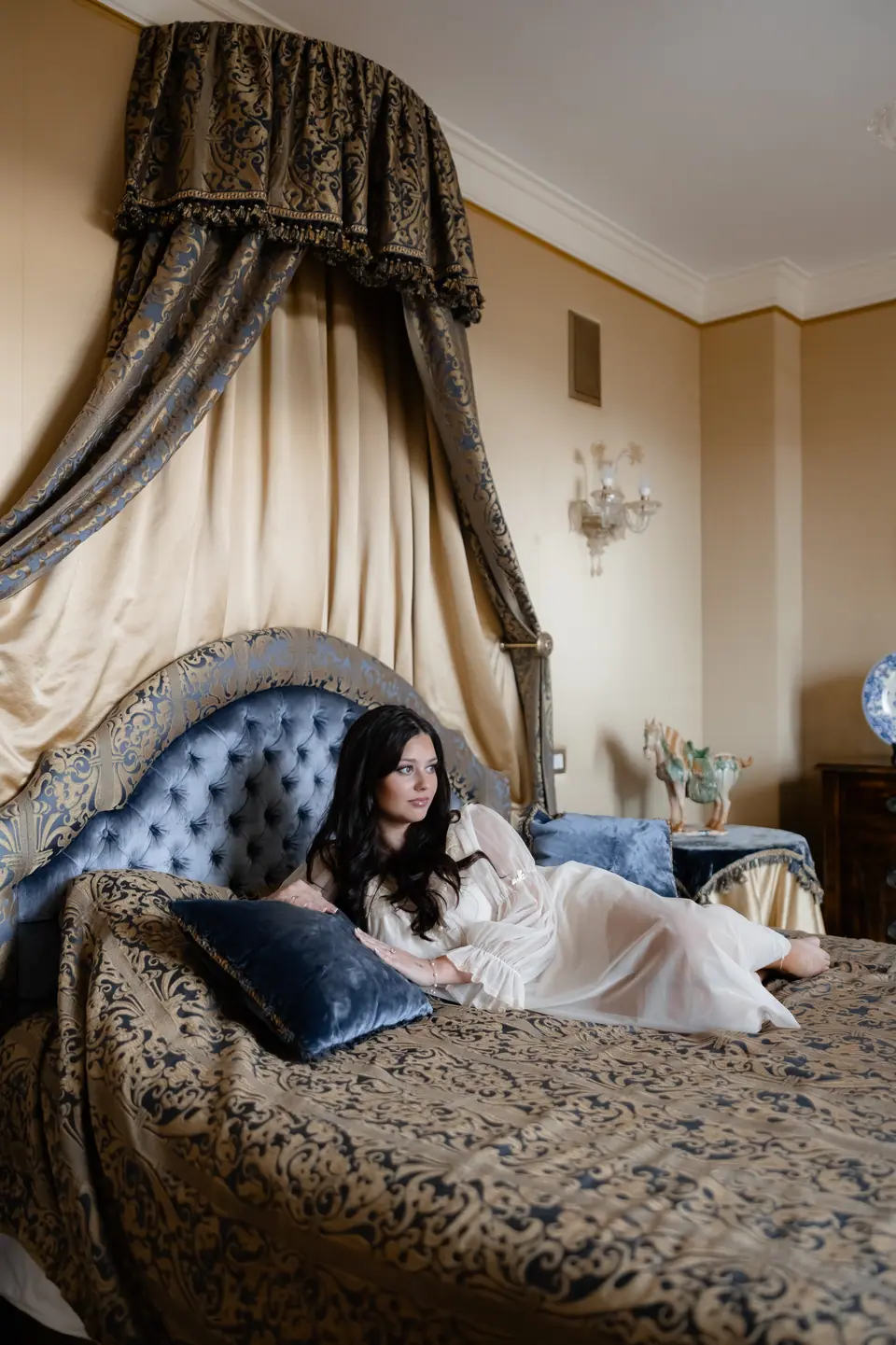 Haley lounging in a sheer white robe on a canopy bed with blue velvet pillows