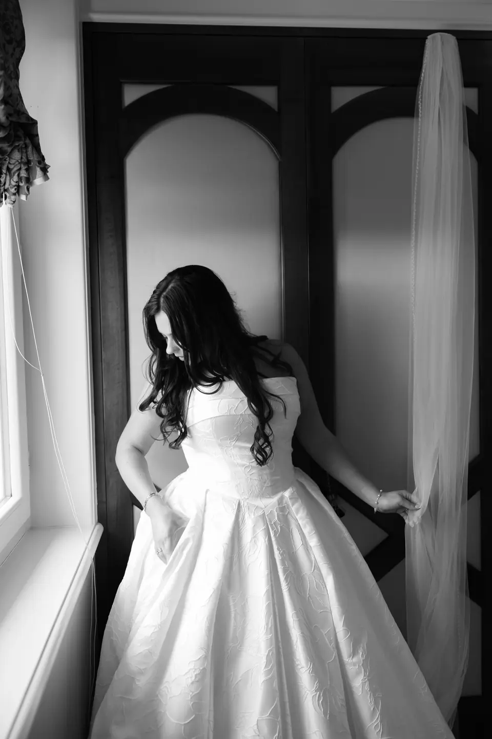 Black and white photograph of Haley in her gown at the window, holding the edge of the curtain