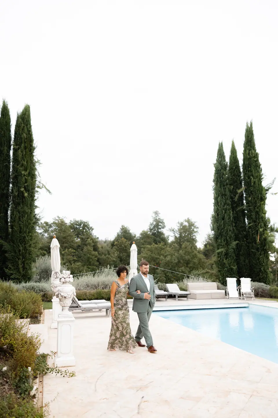 A man in a sage suit walking a woman in a floral dress past the pool