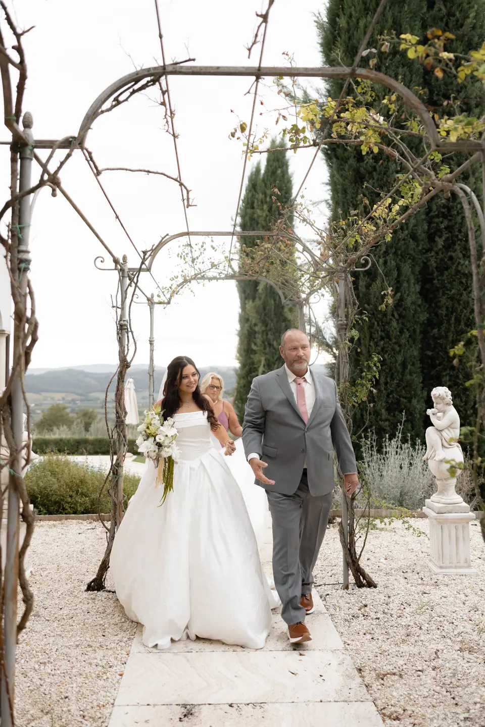 Haley walking down the garden aisle beneath an iron pergola, her arm through a man’s in grey