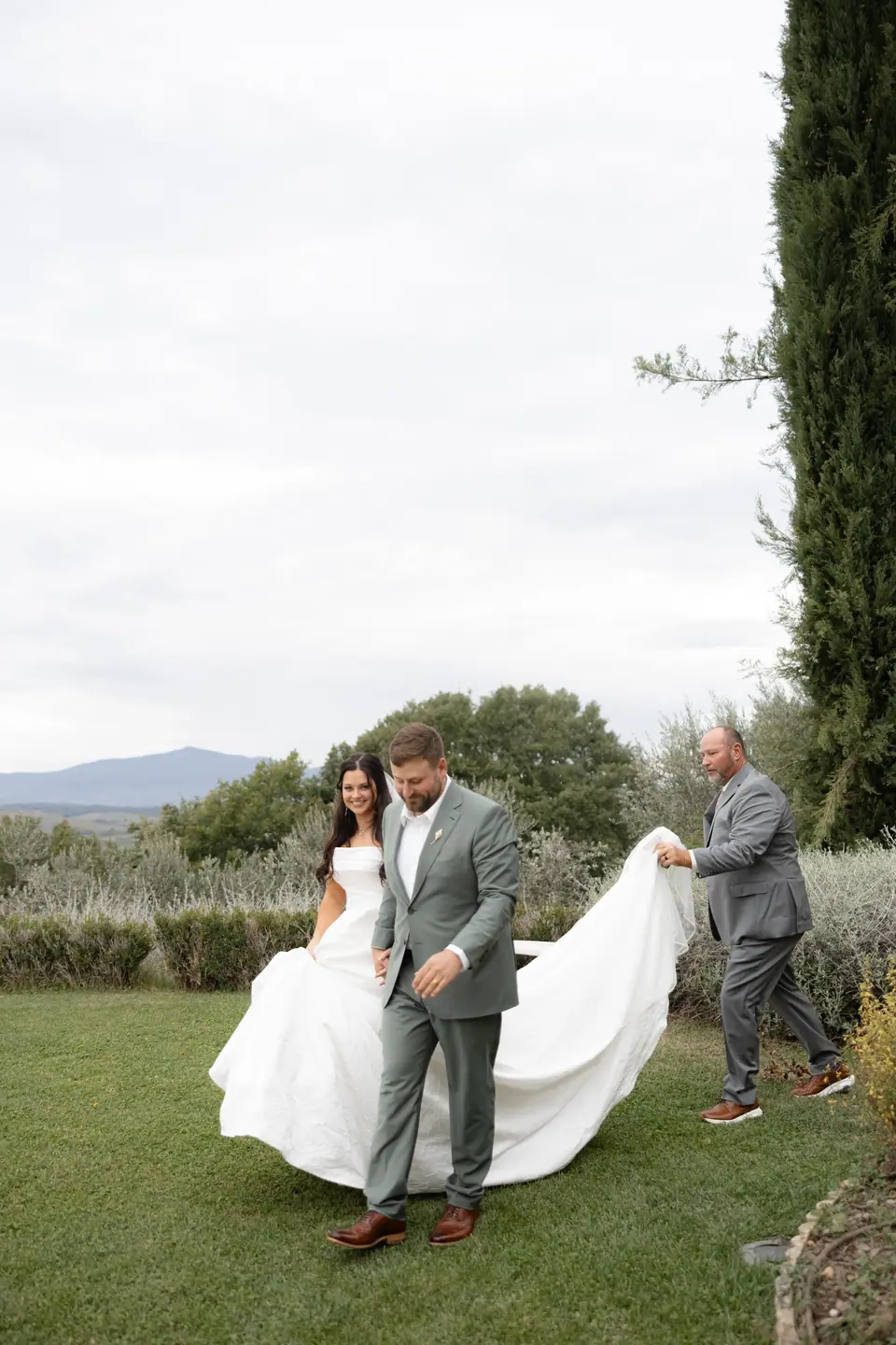 Zach leading Haley across the lawn as a man carries her long train