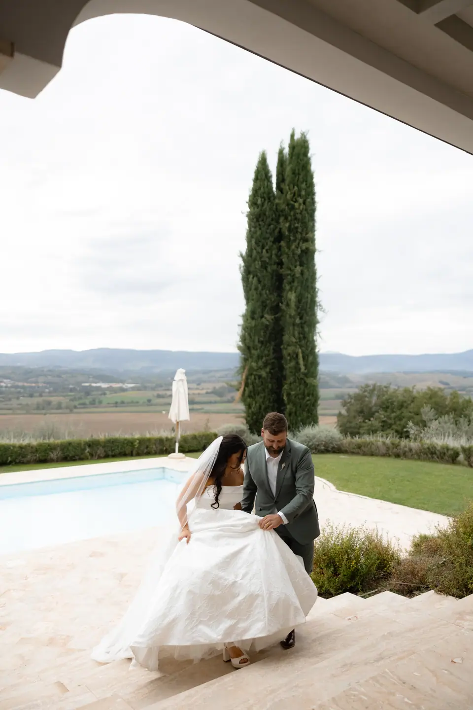 Haley lifting her skirt to climb the pool steps with Zach, a cypress and hills behind