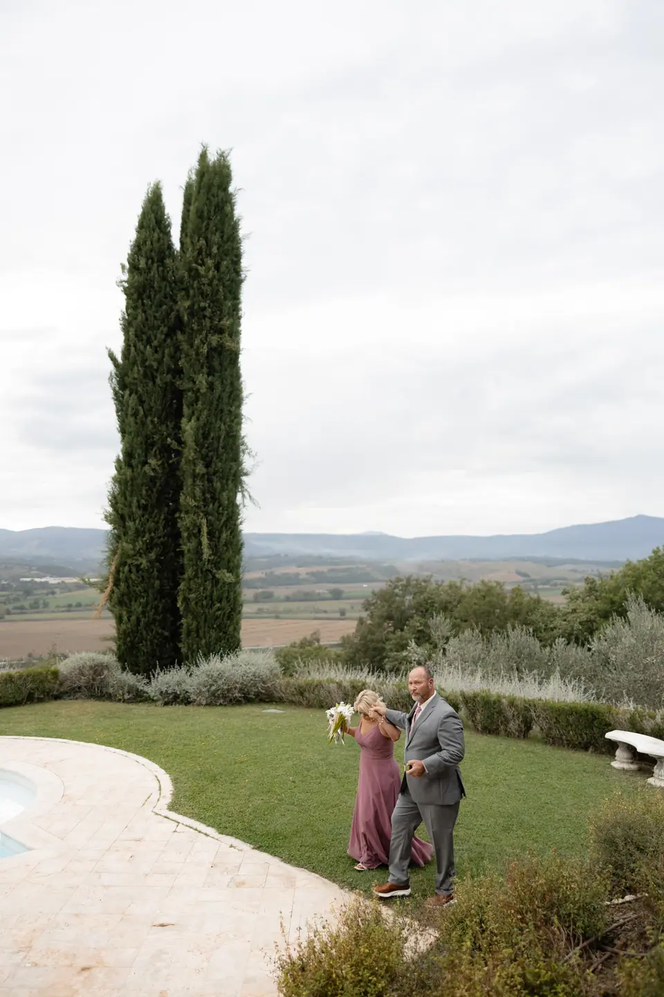 A woman in mauve and a man in grey crossing the lawn below two cypresses
