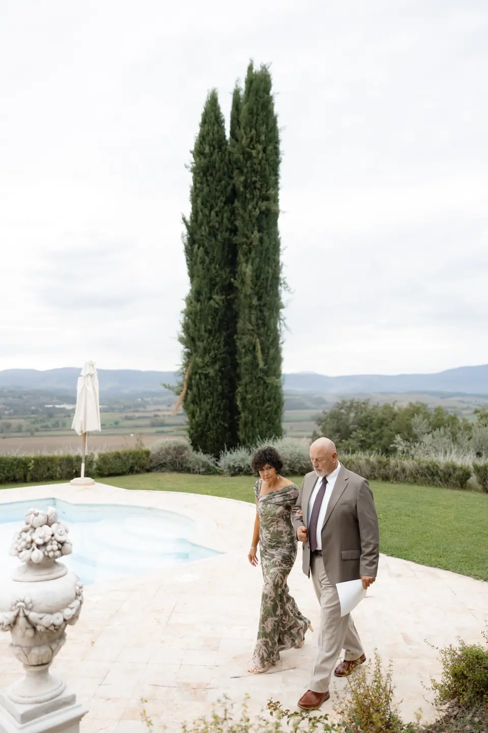 A woman in a floral dress and a man in a tan suit walking along the pool path