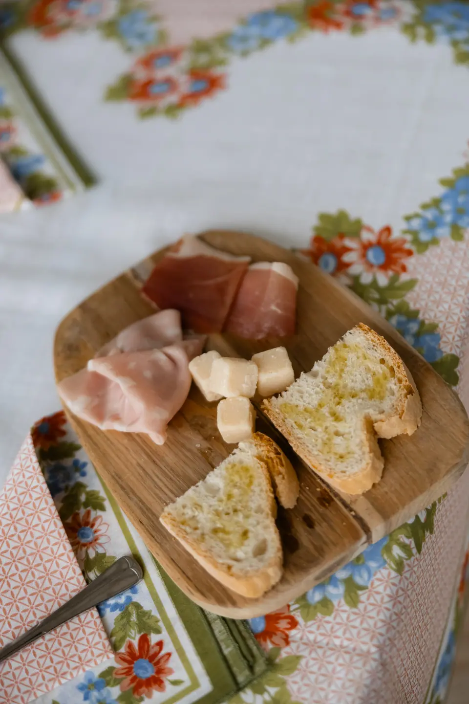 A board of prosciutto, cheese and toasted bread on a floral tablecloth