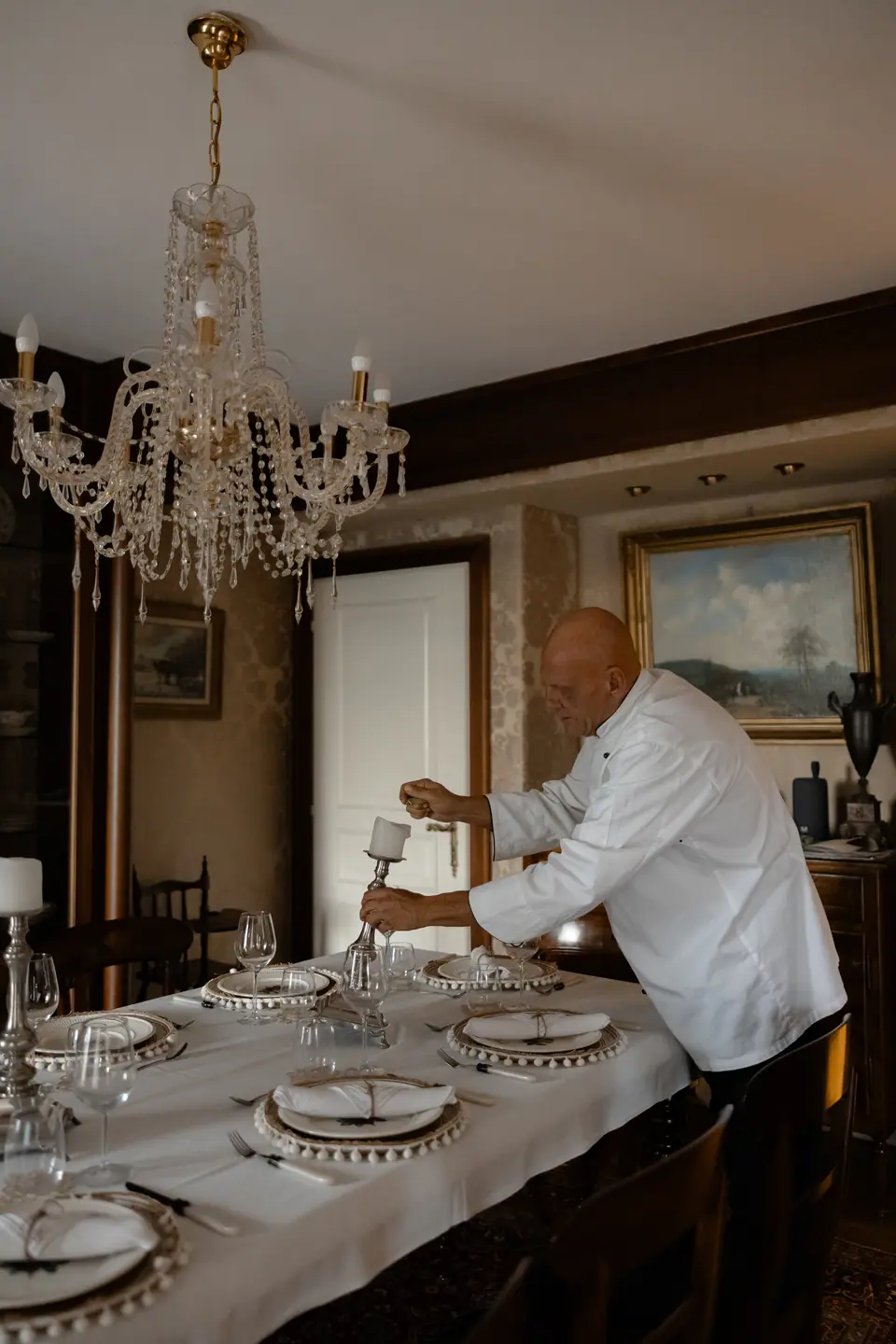 The chef lighting candles at a long dinner table under a crystal chandelier