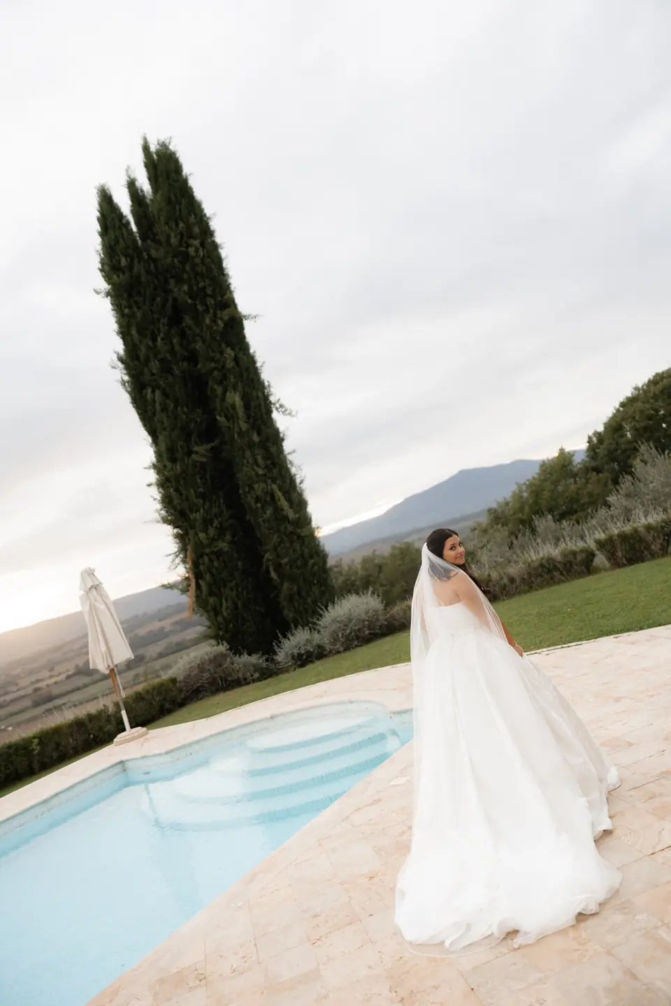 Haley in her gown and veil by the pool, a tall cypress and the valley behind