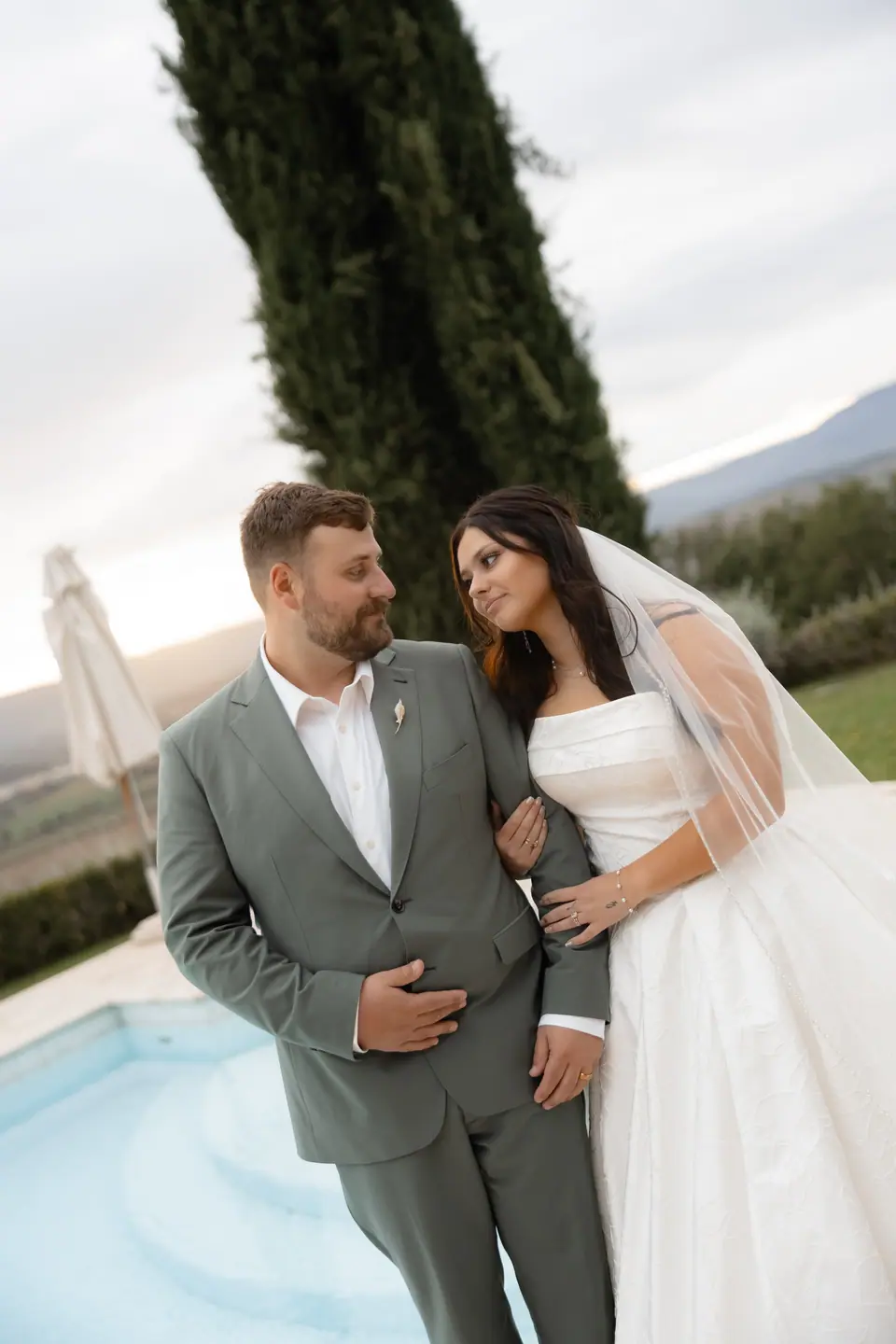 Zach and Haley arm in arm beside the pool, with cypress trees and hills behind