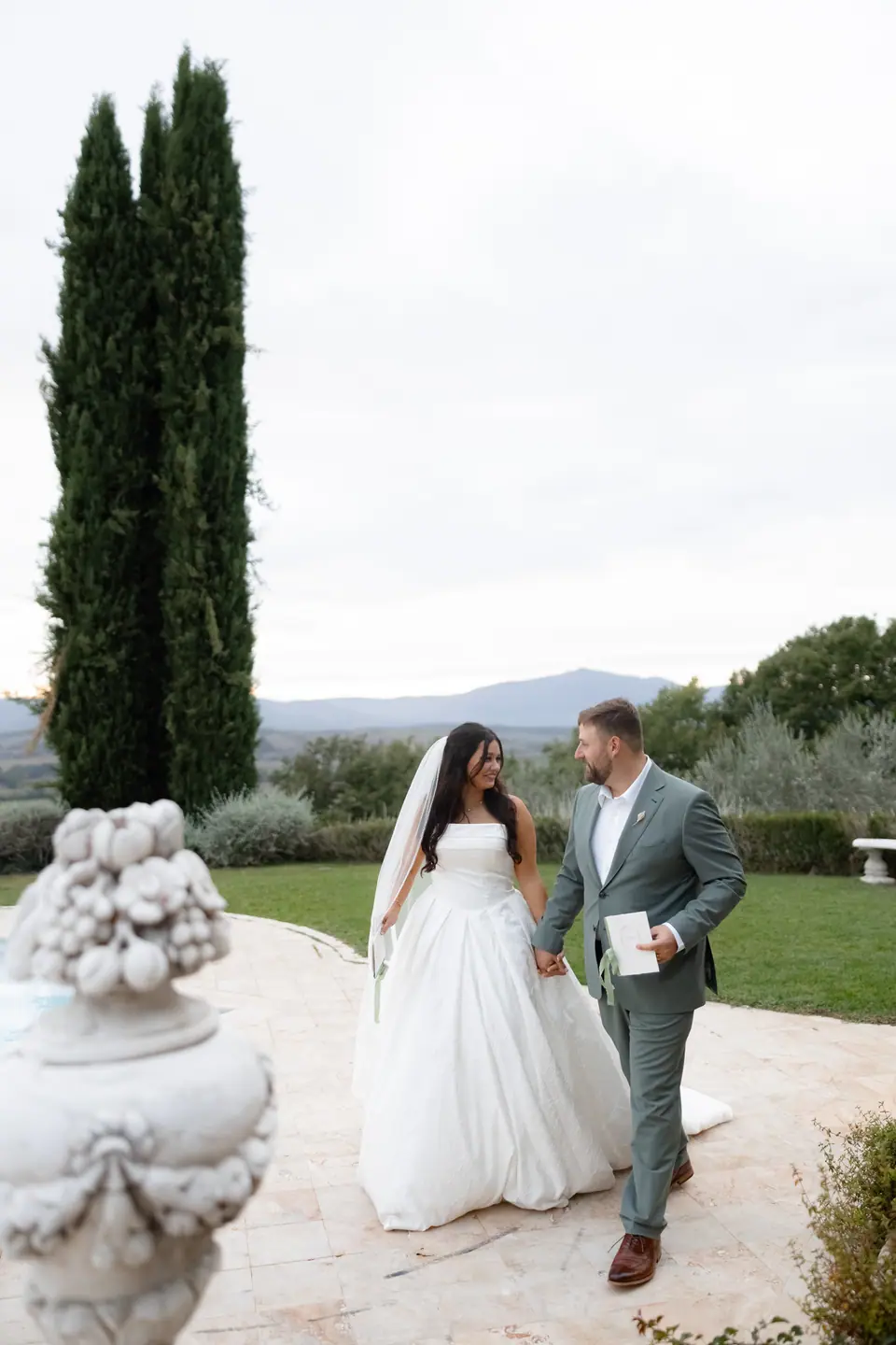 Haley and Zach walking past a stone urn by the pool, letters in hand