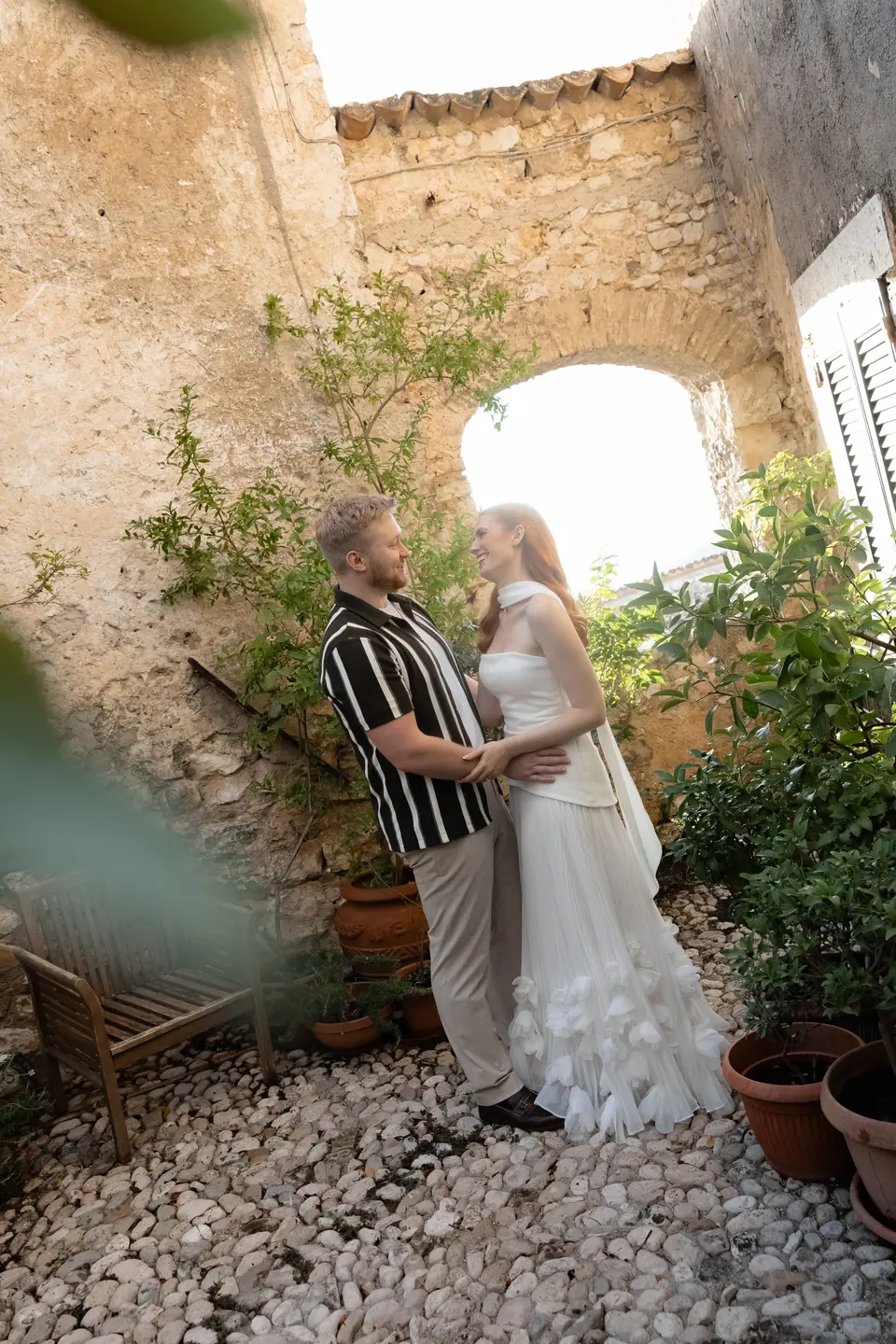 Kate and James embracing in a courtyard beneath a stone arch, among potted plants