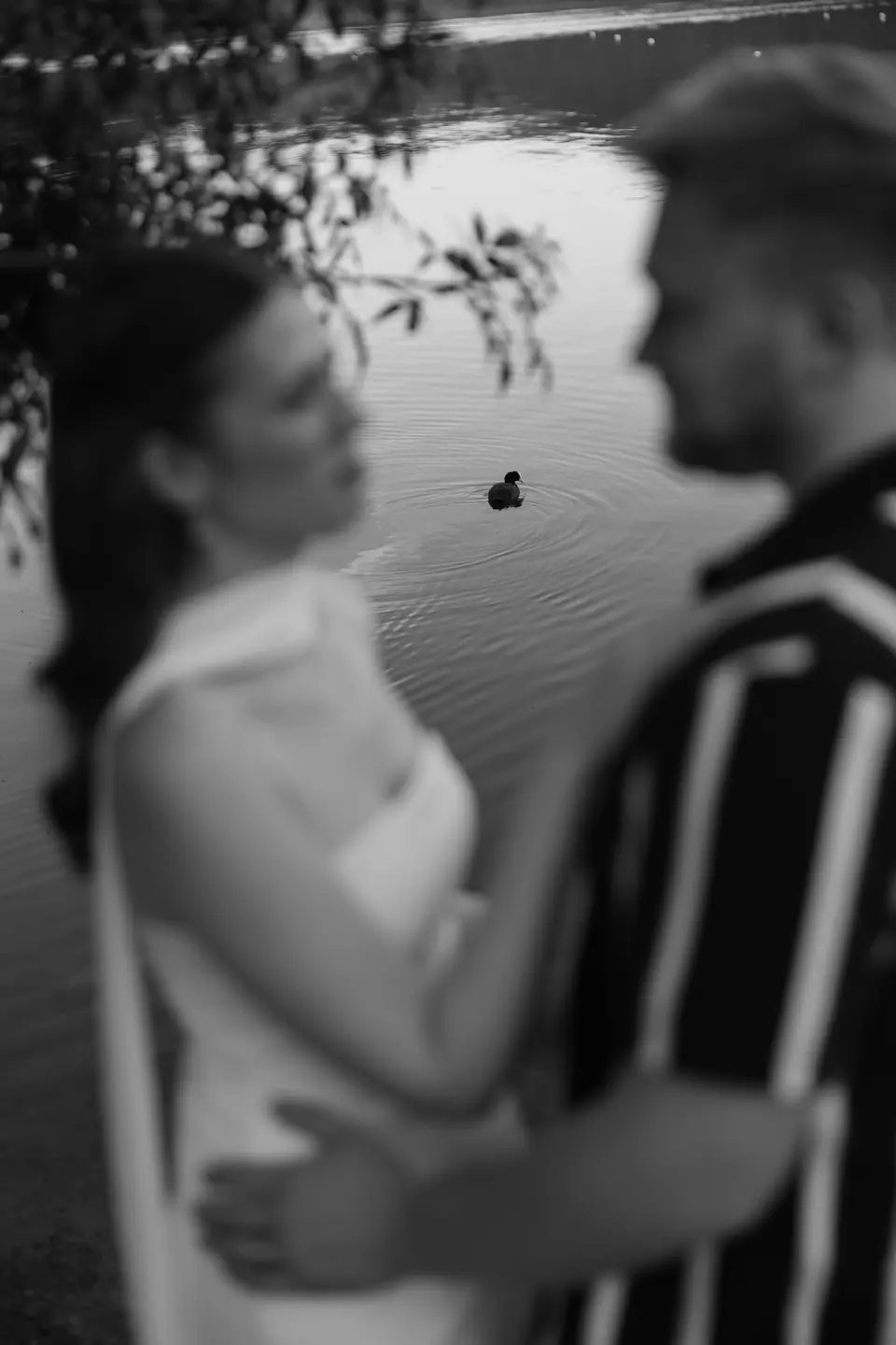 Black and white of Kate and James face to face, a duck on the lake behind them